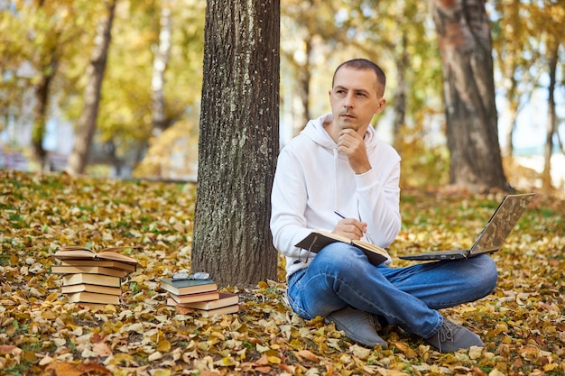 Adult man in a white hoodie is studying in the Park on a laptop writing in a notebook reading books and textbooks Outdoor learning social distancing