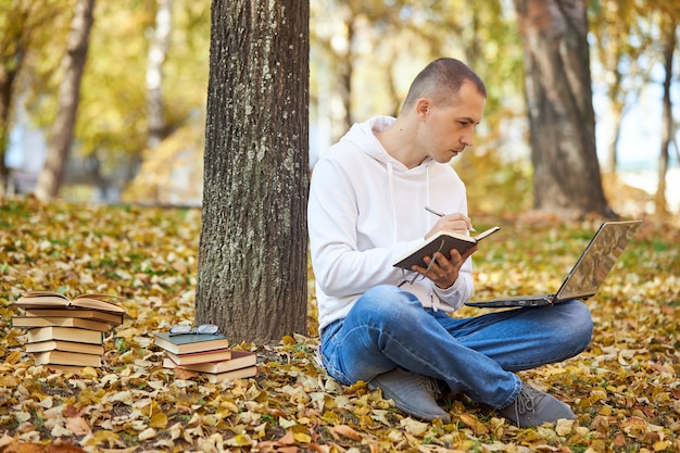 Adult man in a white hoodie is studying in the Park on a laptop, writing in a notebook, reading books and textbooks