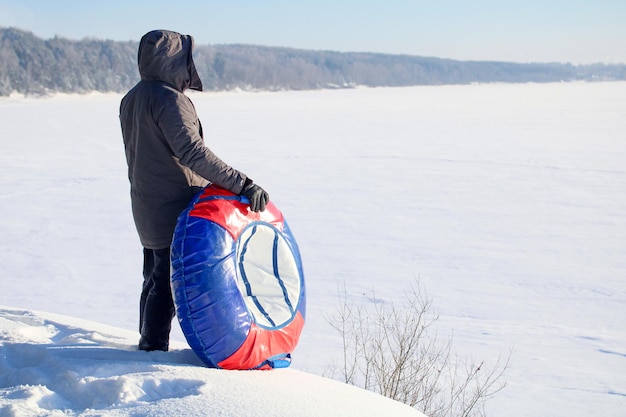 An adult man with a winter inflatable tube stands on the slope by the river