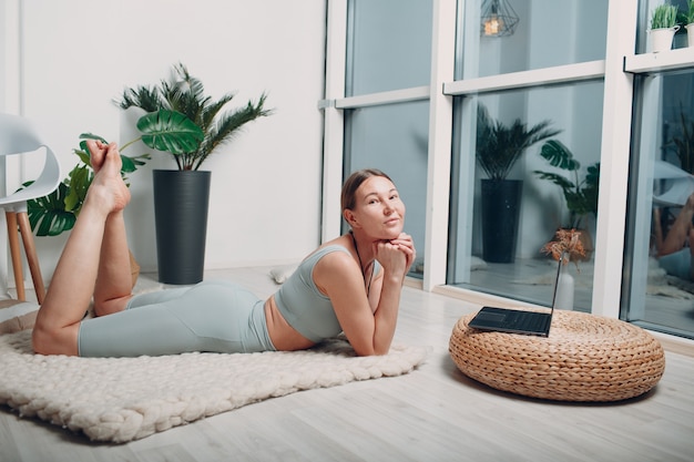 Adult mature woman doing yoga and relaxing at home living room with online tutorials on laptop