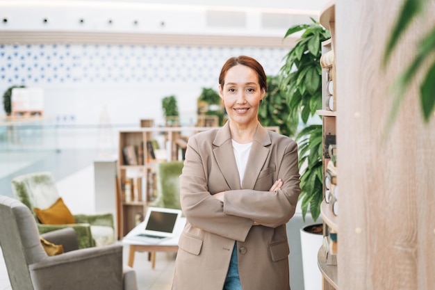 Adult smiling brunette business woman forty years with long hair in stylish beige suit and jeans at public place green open space office coworking Friendly teacher or mentor with book in library