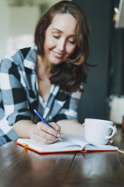 Adult smiling brunette woman in casual doing notes in daily book with cup of tea on kitchen at home