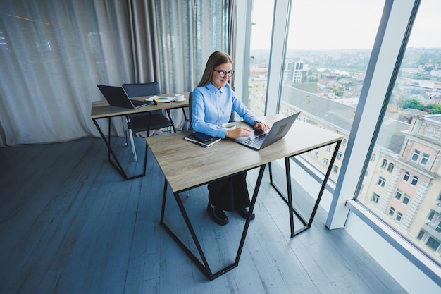 Adult smiling female manager in glasses and formal shirt sits at a desk and looks at a laptop Modern spacious bright office with a large window