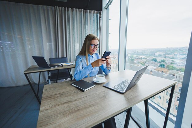 Adult smiling female manager in glasses and formal shirt sits at a desk and looks at a laptop Modern spacious bright office with a large window