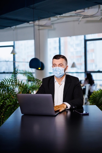 Adult successful handsome male businessman in a mask with a laptop while working in a remote office in a cafe