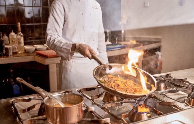 Adult in white chef uniform shaking a pan with pepper slices catching fire inside it over cooker