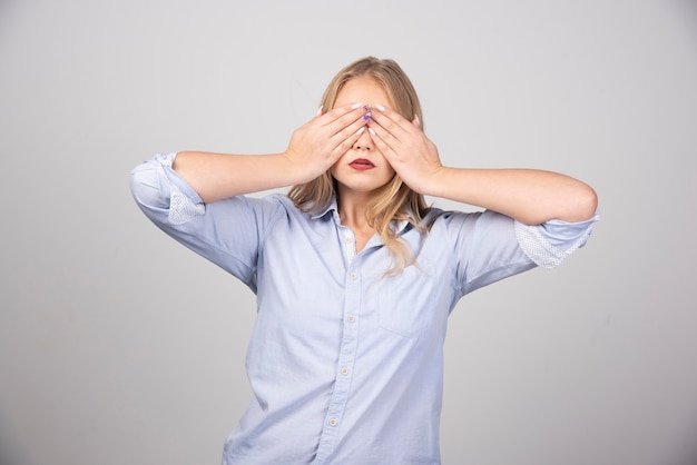 Adult woman covering eyes with hands over gray wall.