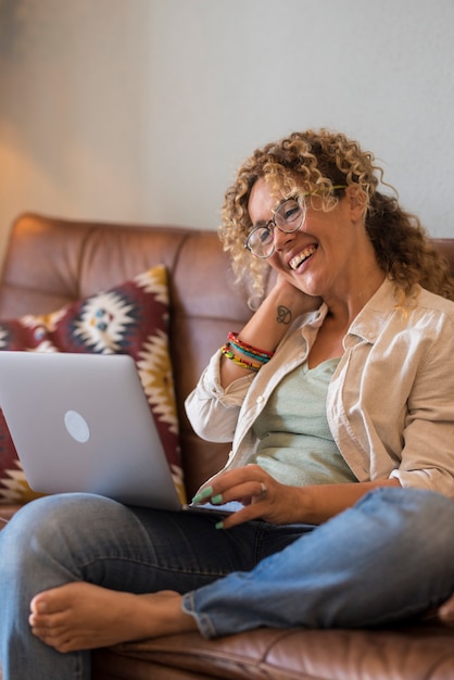 Adult woman sit down on the sofa at home enjoy video call conference with friends smiling and speaking on laptop computer - online people and wireless connection