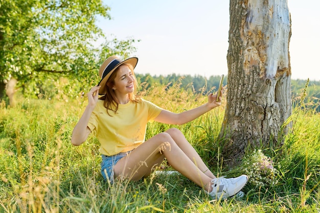 Adult woman in straw hat relaxing sitting on grass looking in smartphone webcam making video call