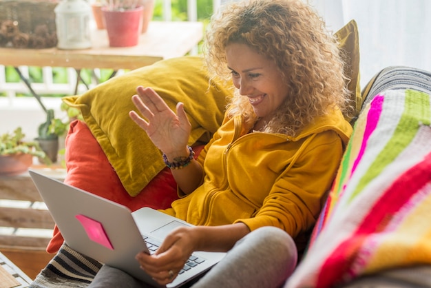 Adult woman in video call use laptop computer sitting on the sofa at home smiling and speaking with friends or parents - wireless connection communication technology and modern people indoor