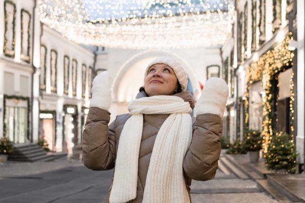 An adult woman in winter clothes in the city looks at the Christmas illumination and admires what she sees.