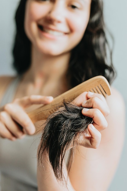 Adult young woman holding wooden hair brush and pulling healthy long black hair isolated on grey Positive charming joy girl getting ready Bodycare effect make hair flawless wellness shine gloss