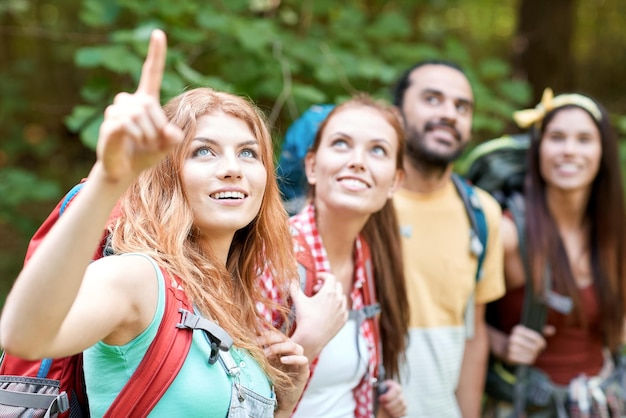 adventure, travel, tourism, hike and people concept - group of smiling friends with backpacks pointing finger in woods