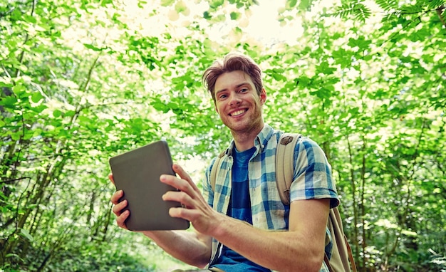 adventure, travel, tourism, hike and people concept - happy young man with backpack and tablet pc computer in woods