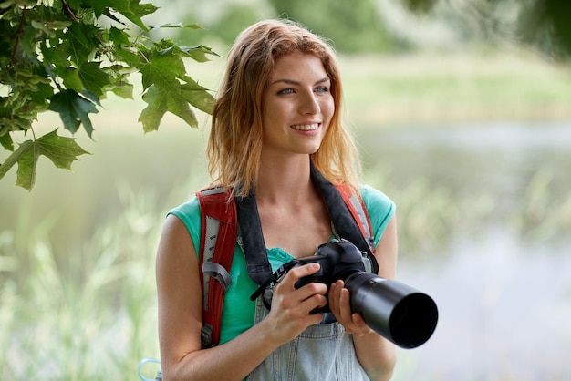 adventure, travel, tourism, hike and people concept - happy young woman with backpack and camera photographing outdoors