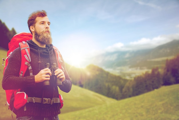 adventure, travel, tourism, hike and people concept - man with red backpack and binocular over alpine mountains and hills background