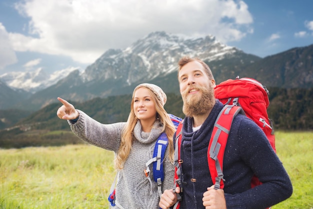 adventure, travel, tourism, hike and people concept - smiling couple walking with backpacks and pointing finger to something over mountains background