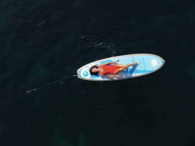 Aerial drone view on well looking middle aged woman with black hair in red swimsuit swimming on sup