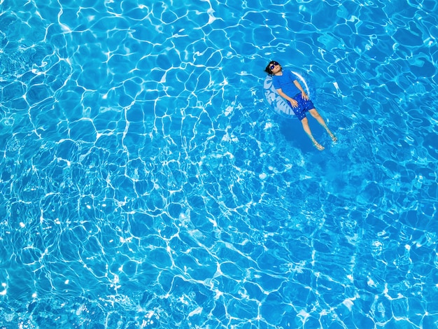 Aerial drone view of  a young boy of ten years old wearing sun protection t-shirt and sunglasses is lying on the inflatable ring in the swimming pool. Summer holidays concept, copy space on the left.