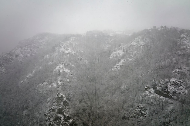 Aerial foggy landscape with mountain cliffs covered with fresh fallen snow during heavy snowfall in winter mountain forest on cold quiet day