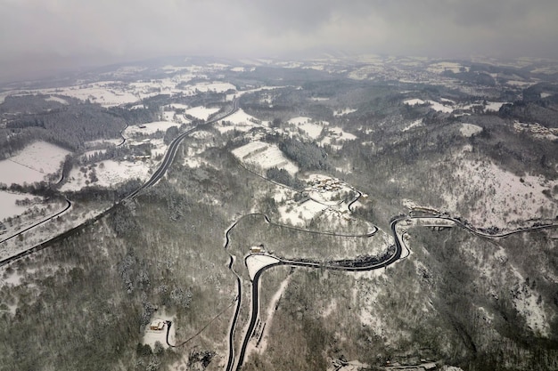 Aerial foggy landscape with mountain road covered with fresh fallen snow during heavy snowfall in winter mountain forest on cold quiet day