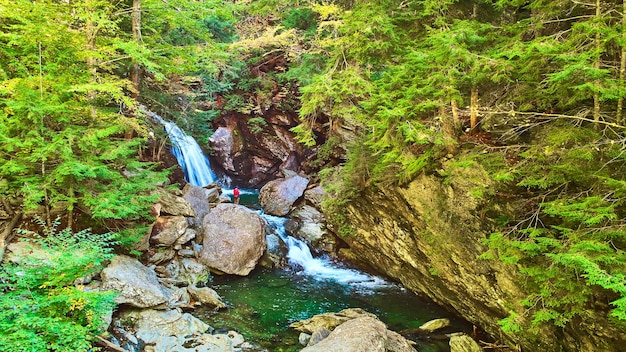 Aerial over river in rocky gorge of lush green forest with waterfall and lone hiker observing falls