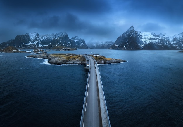 Photo aerial view of bridge island with rorbu sea snowy mountains in fog blue cloudy sky at twilight in spring dramatic landscape top drone view of road hamnoy village lofoten islands norway