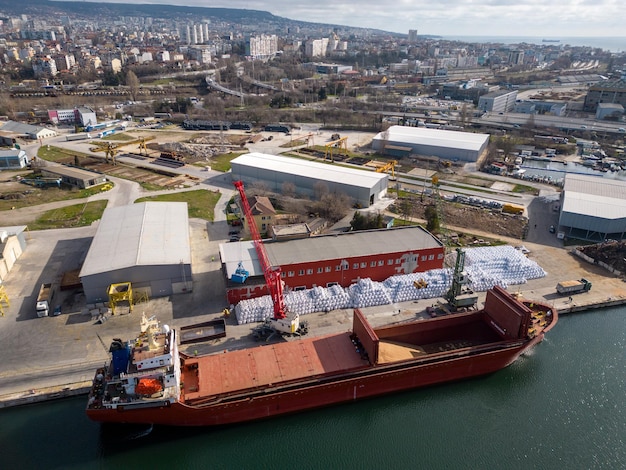 Aerial view of a bulk carrier ship loaded with grain is docked at a busy port with workers and machinery seen in the background