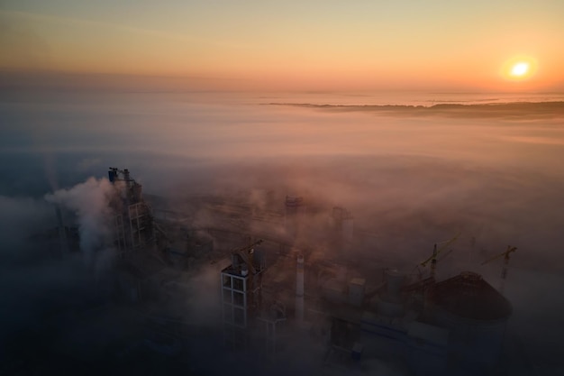 Aerial view of cement factory with high concrete plant structure and tower crane at industrial production site on foggy morning. Manufacture and global industry concept.