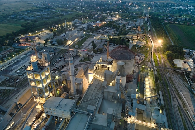 Aerial view of cement factory with high concrete plant structure and tower cranes at industrial production area at night. Manufacture and global industry concept.