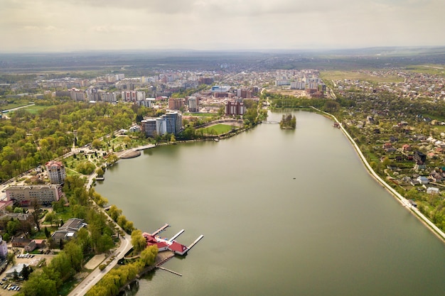 Aerial view of city lake among green trees and town buildings in recreation park zone. Drone photography.