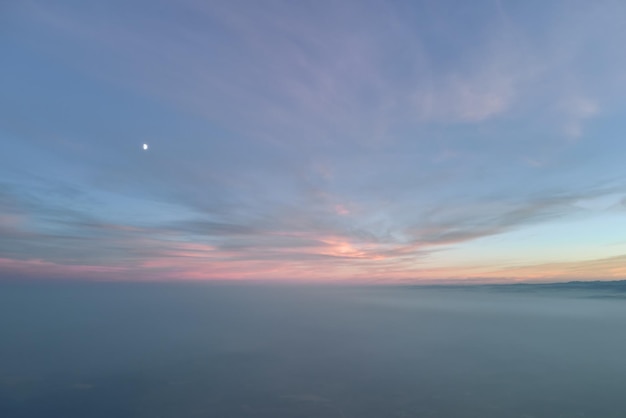 Aerial view of colorful sunset over white dense foggy clouds cover with distant dark silhouettes of mountain hills on horizon