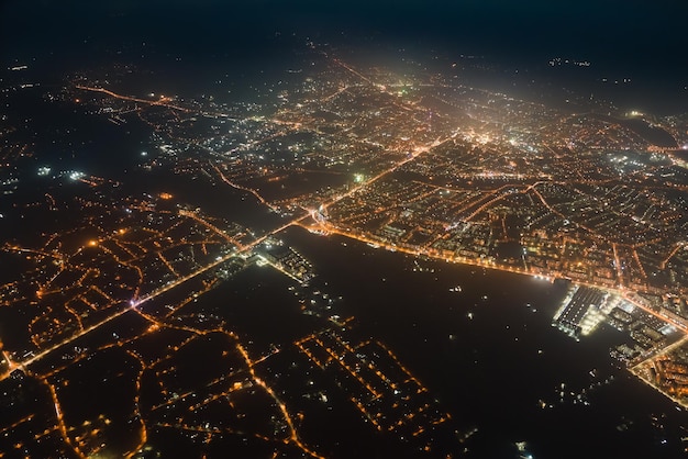 Aerial view from airplane window of buildings and bright illuminated streets in city residential area at night. Dark urban landscape at high altitude