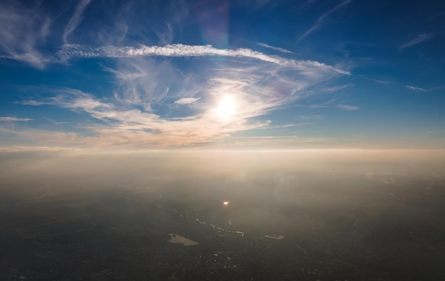 Aerial view from airplane window at high altitude of distant city covered with layer of thin misty smog and distant clouds in evening