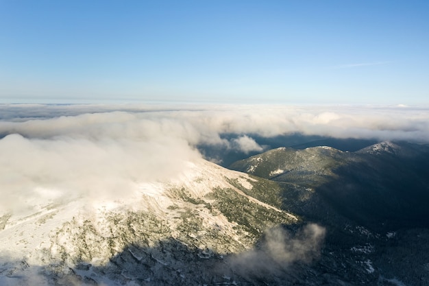 Aerial view from above of white puffy clouds covering snowy mountain tops in bright sunny day.