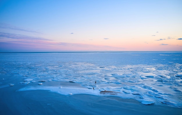 Aerial view two people walk on ice on sunset over the frozen sea winter landscape on seashore during