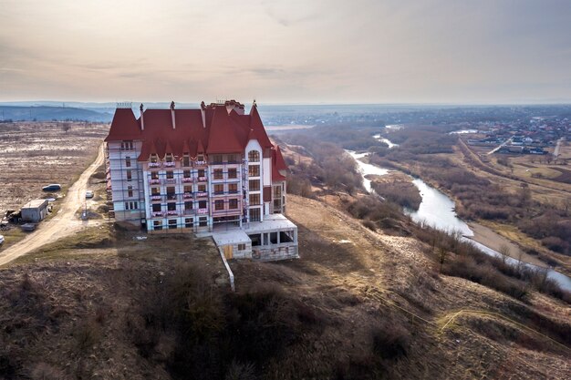 Aerial view of unfinished multistoried residence, hotel or cottage building with stucco wall, cast iron balcony railings, steep shingle roof and shiny windows on rural landscape background.