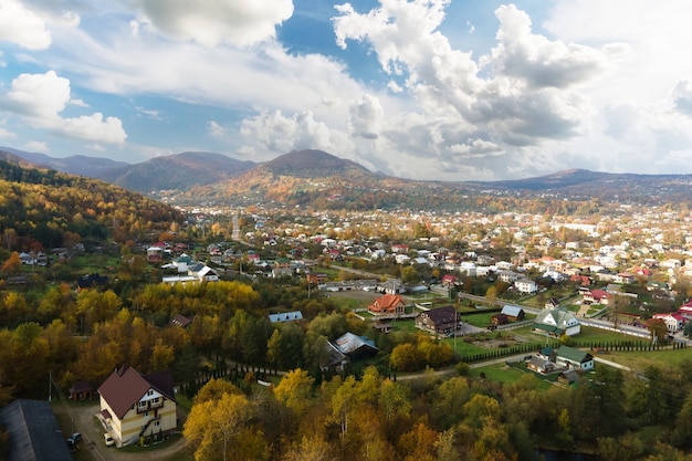 Aerial view of a village rural area with small houses between autumn mountain hills covered with yellow and green spruce forest.