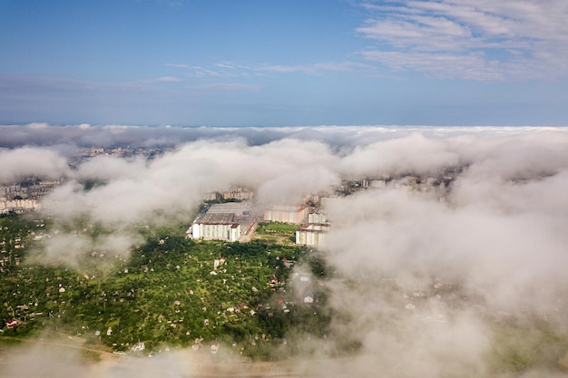 Aerial view of white clouds above a town or village with rows of buildings and curvy streets between green fields in summer Countryside landscape from above