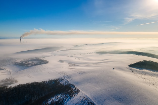 Aerial view of winter landscape with foggy countryside and distant factory pipes emmiting black dirty smoke polluting environment.