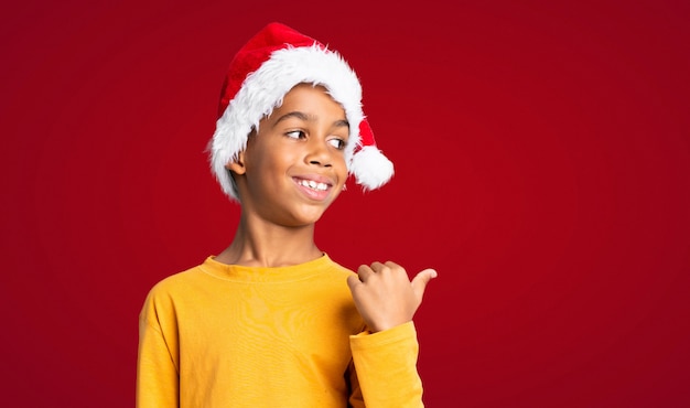 African American boy with christmas hat pointing to the side to present a product over red wall