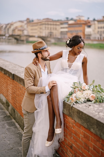 An African-American bride sits on a brick wall and a Caucasian groom hugs her. The embankment of the Arno River, overlooking city and bridges. Interracial wedding couple