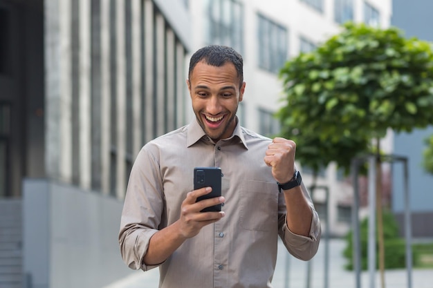 African american businessman outside office building using phone smiling and happy holding hand up