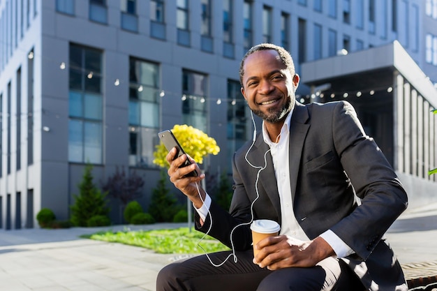 African american businessman sitting outside on a bench near an office center wearing headphones