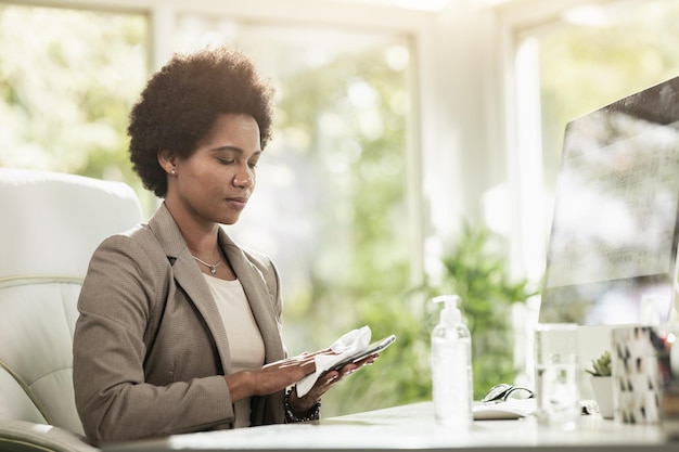 African American businesswoman using antiseptic gel to disinfect her smartphone while sitting in an office alone at work.