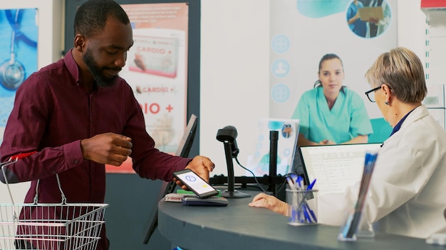 African american client using mobile nfc payment on pos to pay for vitamins and pills, buying prescription treatment at drugstore desk. Adult at cash register paying for supplements. Handheld shot.