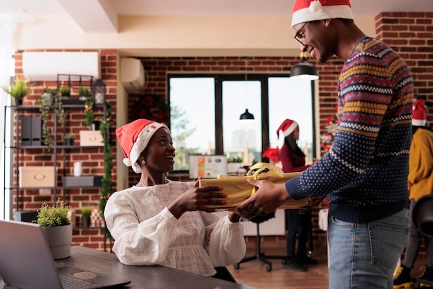 African american colleagues exchanging xmas gift box in festive office with christmas tree to celebrate winter holiday. People giving seasonal presents and feeling cheerful about santa.