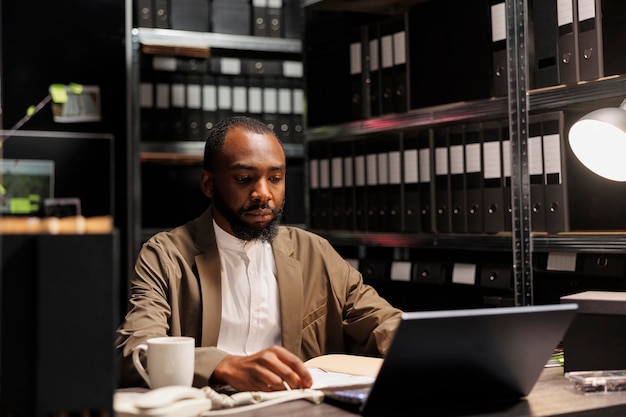 African american cop working overtime on laptop in dark detective office. Serious investigator analyzing police report and searching insight for solving crime at night time