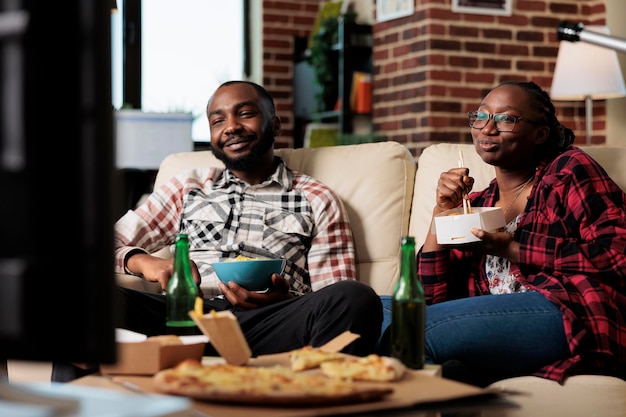 African american couple eating takeaway meal with noodles and chips to watch movie on television. Leisure activity together with film on tv and fast food delivery package, having fun.