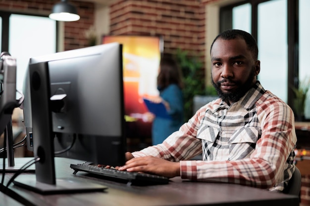 African american creative company employee sitting at desk in office workspace while looking at camera. Agency adult worker at computer in startup studio space modern interior.